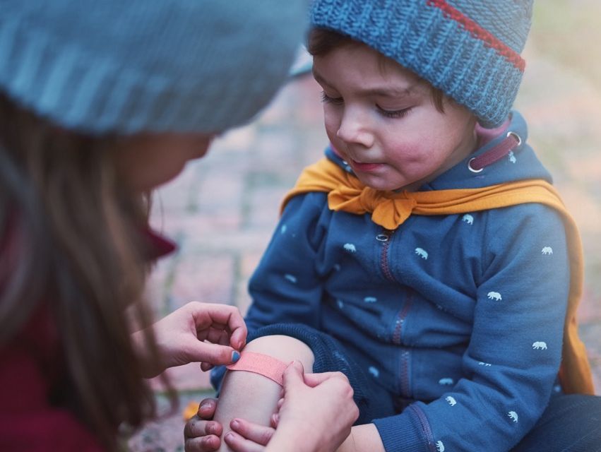 zur Vergrößerungsansicht des Bildes: Kleinkind sitzt mit Gummistiefeln am Boden und bekommt Pflaster aufs Knie.